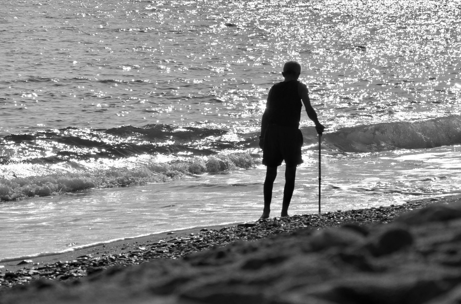 Un anziano che passeggia lungo la spiaggia al controluce, in bianco e nero